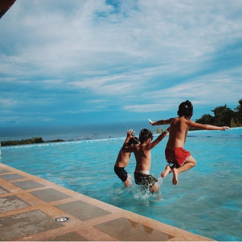 Tre bambini si tuffano insieme in una piscina all’aperto, circondati da un panorama mozzafiato che guarda il mare. L’immagine trasmette il valore di un ambiente sicuro e curato, dove la copertura automatica elettrica della piscina non è solo una questione di efficienza, ma anche di protezione per tutta la famiglia. 