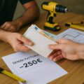 Pagamento in contanti senza fattura per lavori in nero in casa. Due mani si scambiano una busta contenente banconote, con attrezzi da cantiere sullo sfondo.