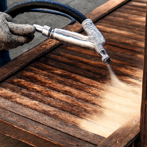 Sabbiatura del legno con getto abrasivo su superficie antica. Operatore che effettua la sabbiatura di una superficie in legno con ugello a pressione, mostrando il contrasto tra legno usurato e pulito.