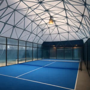 Campo da padel indoor all’interno di una cupola geodetica in acciaio con copertura traslucida e pavimento blu.