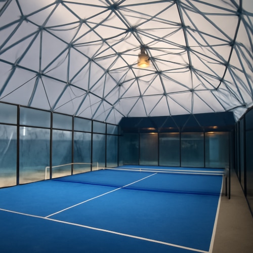 Campo da padel indoor all’interno di una cupola geodetica in acciaio con copertura traslucida e pavimento blu.