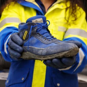 Vecchia scarpa antinfortunistica usata mostrata in primo piano da un’operatrice con giacca ad alta visibilità e guanti blu (divisa di Poste Italiane).