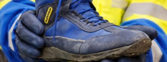 Vecchia scarpa antinfortunistica usata mostrata in primo piano da un’operatrice con giacca ad alta visibilità e guanti blu (divisa di Poste Italiane).