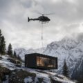Baita prefabbricata di lusso total black trasportata in elicottero e posata su un pendio innevato di montagna.