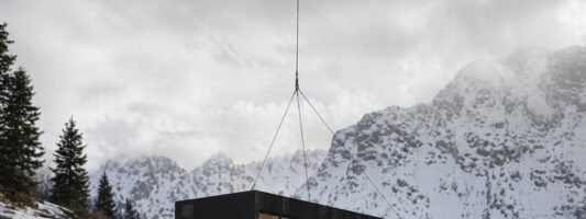 Baita prefabbricata di lusso total black trasportata in elicottero e posata su un pendio innevato di montagna.