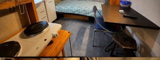 Piccola stanza arredata con letto singolo, piastra elettrica senza gas, scrivania, armadio e TV disposti in uno spazio molto ridotto. L’immagine richiama il caso del monolocale n affitto a Modena e mette in evidenza la compressione estrema degli ambienti domestici.