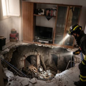 Un vigile del fuoco con casco da soccorso e due luci forti splende una torcia in una massiccia voragine causata dal crollo del pavimento piastrellato di un soggiorno residenziale.